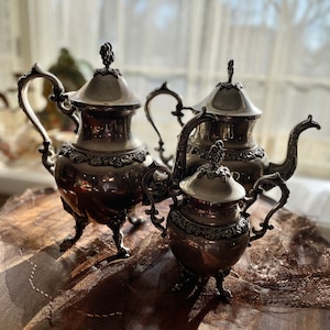 Puede incluir: Un juego de té de plata con tetera, jarra de crema y azucarero. El juego tiene un diseño ornamentado con un patrón floral.