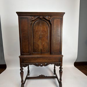May include: A dark brown antique cabinet with ornate carvings. The cabinet has two doors and sits on a stand with four legs. The wood grain is visible throughout the piece. The cabinet is approximately 152 cm tall.