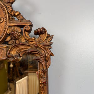 May include: Ornate, antique mirror with a gold-colored, carved wooden frame. The frame features detailed floral and scrollwork designs, with hints of red paint. The mirror reflects a room's interior, adding depth to the image.