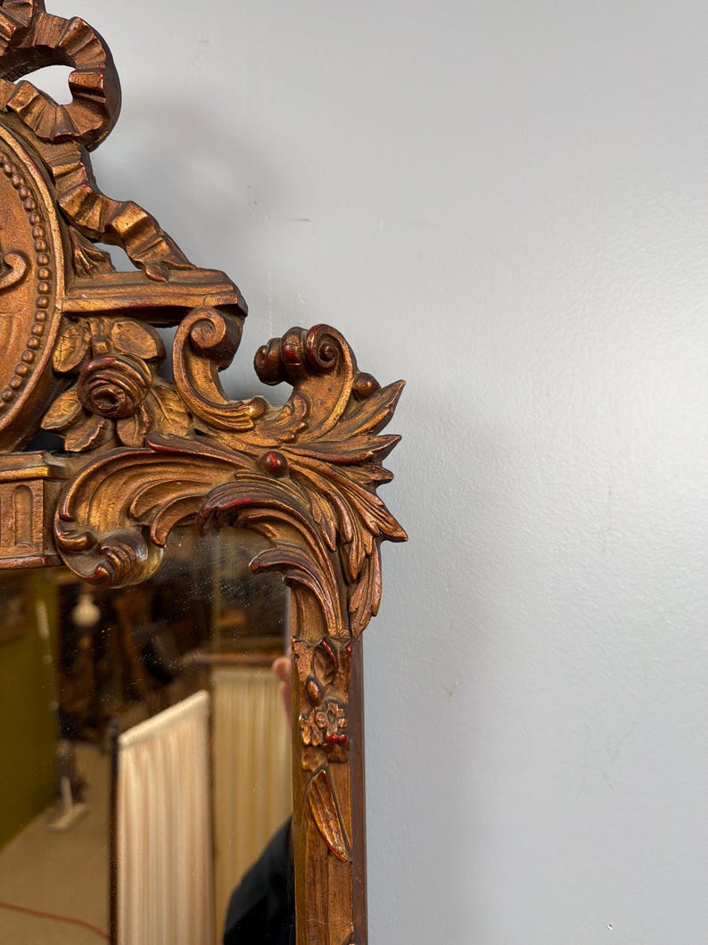 May include: Ornate, antique mirror with a gold-colored, carved wooden frame. The frame features detailed floral and scrollwork designs, with hints of red paint. The mirror reflects a room's interior, adding depth to the image.