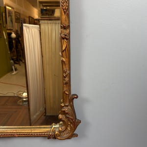 May include: Ornate, rectangular gold-framed mirror. The frame features detailed carvings of floral and leaf motifs, with a decorative scroll design at the corner. The mirror reflects a room with artwork and curtains.