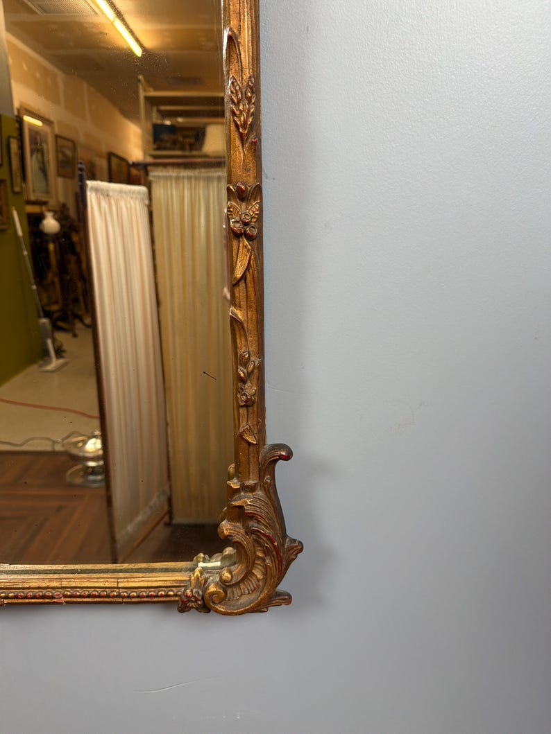 May include: Ornate, rectangular gold-framed mirror. The frame features detailed carvings of floral and leaf motifs, with a decorative scroll design at the corner. The mirror reflects a room with artwork and curtains.