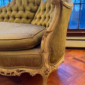 May include: An antique olive green chaise lounge with a tufted back and seat. The wooden frame is painted in a cream color and features ornate carvings. The chaise lounge is placed in front of a window.
