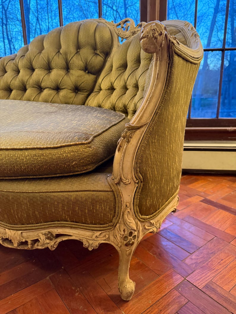 May include: An antique olive green chaise lounge with a tufted back and seat. The wooden frame is painted in a cream color and features ornate carvings. The chaise lounge is placed in front of a window.