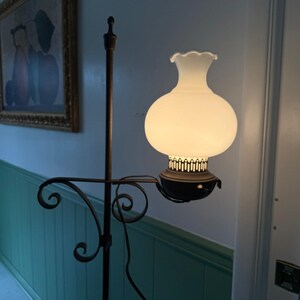 May include: A vintage floor lamp with a white glass globe shade and a dark metal frame. The lamp features a decorative, scrolled arm and a tall, slender pole. The shade has a ruffled top, and the lamp is plugged into an electrical outlet.
