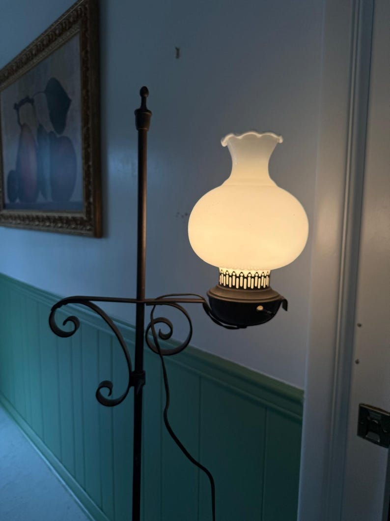 May include: A vintage floor lamp with a white glass globe shade and a dark metal frame. The lamp features a decorative, scrolled arm and a tall, slender pole. The shade has a ruffled top, and the lamp is plugged into an electrical outlet.