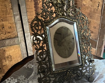 May include: Ornate antique brass picture frame with a detailed, scrolling design. The frame features a central oval opening with a mirrored surface and a decorative face at the top. The frame is displayed on a white lace cloth.