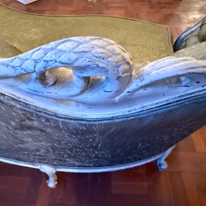 May include: Antique chaise lounge with a carved, white-painted wooden armrest. The chaise has a dark, patterned seat and a curved back. The legs are also white and have a decorative design. The overall style is vintage and elegant.