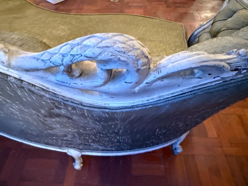 May include: Antique chaise lounge with a carved, white-painted wooden armrest. The chaise has a dark, patterned seat and a curved back. The legs are also white and have a decorative design. The overall style is vintage and elegant.