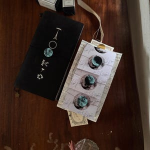 Op de afbeelding: Een zwart notitieboek met een maanfase-ontwerp, naast kleine doosjes en decoratieve stickers. Het notitieboek heeft een gestileerde Eiffeltoren en bloemmotieven. De stickers tonen een halve maan met een turquoise en blauw ontwerp.