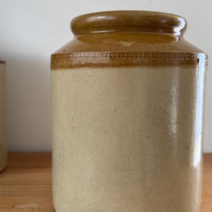 May include: A classic stoneware jar, featuring a light beige body and a brown, glazed top. The jar has a rounded form and a wide opening. The top is adorned with a decorative band. Likely used for storage, it's a vintage piece.