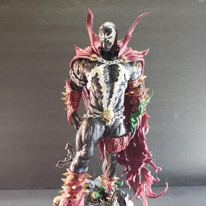 May include: A detailed figurine of the comic book character Spawn, standing on a rocky base. The figure is predominantly black with white accents, a red cape, and spiked armor. A silver chain with a skull hangs around the waist.