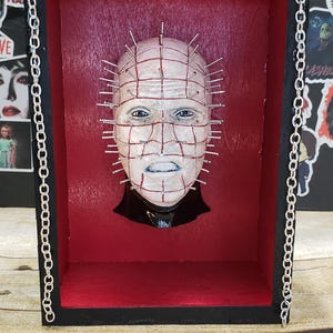 May include: A horror-themed shadow box featuring a sculpted face with numerous metal pins. The face is set against a red background within a black frame, with a silver chain on the side. The artwork is a unique piece of horror decor.