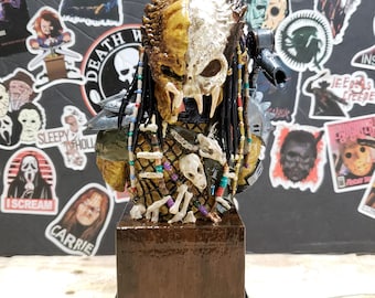 May include: A detailed bust of a Predator character with a skull-like mask, painted in a mix of brown, white, and green. The bust is adorned with beads and bones, and is displayed on a wooden base with a black trim.