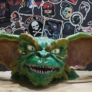 May include: A green and brown detailed figurine of Gizmo from the movie Gremlins. The figurine has large ears and a toothy grin. The figurine is sitting on a wooden surface.