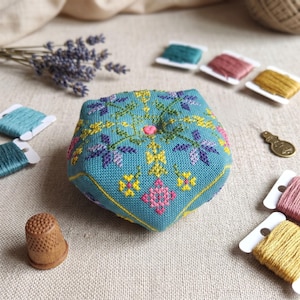 Peut inclure: Un coussin à épingles turquoise avec broderies florales jaunes, roses et violettes. Autour du coussin se trouvent des bobines de fil de différentes couleurs, un dé à coudre en bois et une branche de lavande. Une petite épingle en forme de cœur est au centre.