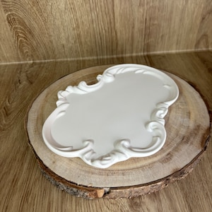 Peut inclure: Plaque en céramique blanche avec une bordure décorative et ornée. La plaque est posée sur une tranche de bois.