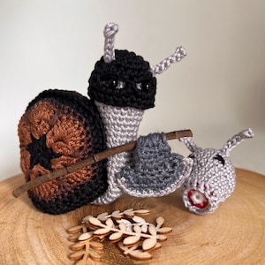May include: A handmade crochet snail figurine with a black and brown shell, grey body, and a small wooden stick. The snail appears to be pulling a smaller grey crochet donkey figurine. A small pile of wooden leaf cutouts is in front.