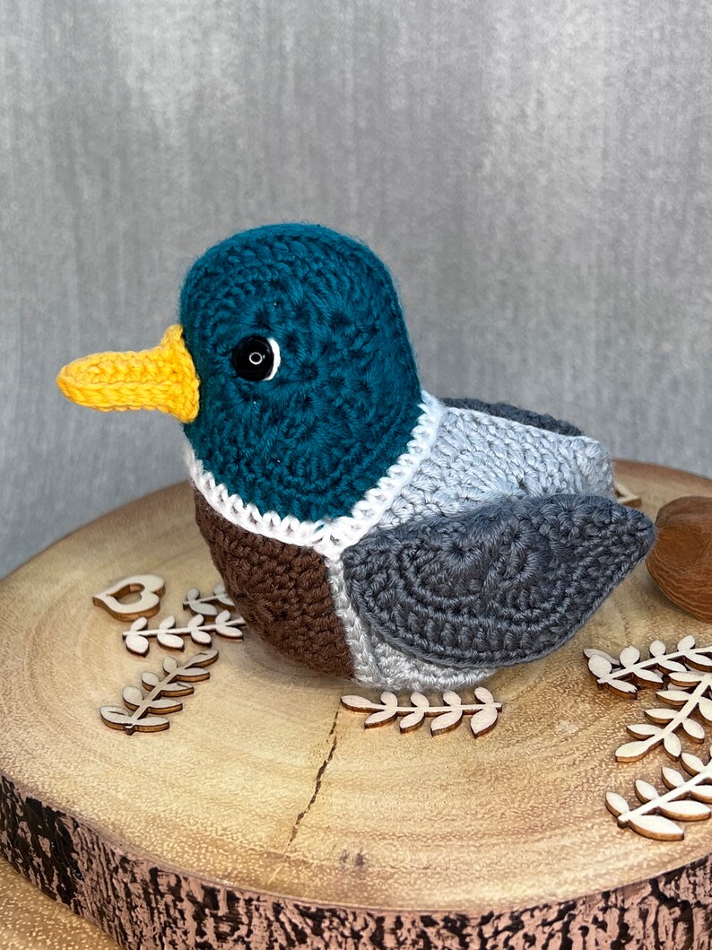 May include: A crocheted duck toy with a teal head, brown chest, grey wings, and a yellow beak. The duck is sitting on a wooden surface with decorative wooden leaves.