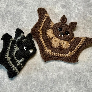 Può includere: Due ornamenti a forma di pipistrello all'uncinetto. Uno è nero con strisce grigie, l'altro è marrone con accenti beige e marrone scuro. Entrambi hanno zanne bianche e occhi neri. Perfetti per le decorazioni di Halloween.