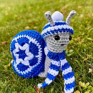 Puede incluir: Un caracol de crochet con un gorro y una bufanda a rayas azules y blancas. El caracol está sentado en un lecho de hierba verde.