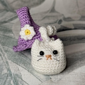 May include: A white crocheted cat with black eyes and a brown nose wearing a purple crocheted hat with a white flower.