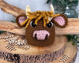 May include: A brown crocheted Highland cow with pink ears, black eyes, and a golden brown mane. The cow is sitting on a wooden log.