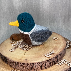 May include: A crocheted duck toy with a teal head, brown body, and gray wings. The duck is sitting on a wooden log slice with decorative wooden leaves.