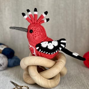May include: A crocheted red and black bird with a long beak and a white and black patterned wing. The bird is sitting on a wooden knot.