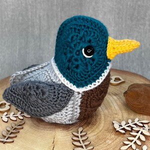 May include: A crocheted duck plush toy with a teal head, grey wings, brown chest, and a yellow beak. The duck is sitting on a wooden surface with wooden leaf decorations.