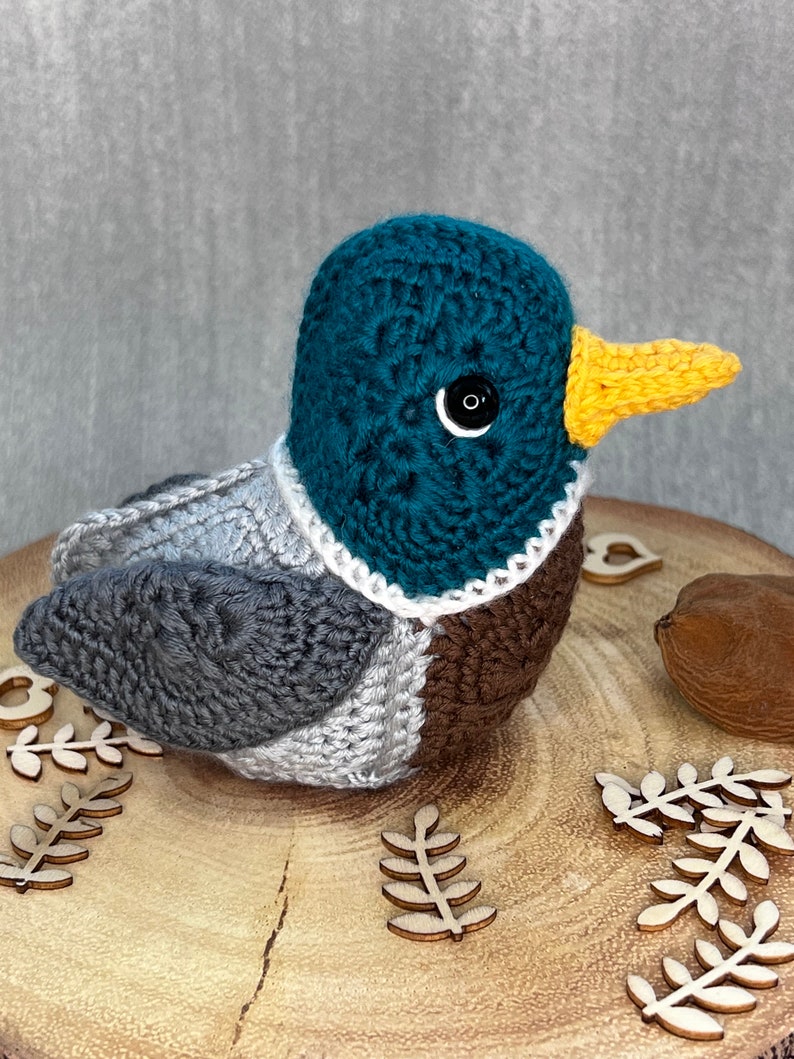 May include: A crocheted duck plush toy with a teal head, grey wings, brown chest, and a yellow beak. The duck is sitting on a wooden surface with wooden leaf decorations.