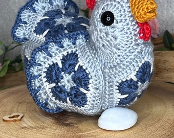 May include: A handmade crochet chicken in shades of gray and navy blue, with a red comb and yellow beak. The chicken sits on a wooden slice next to a white egg. The crochet work features a detailed pattern.