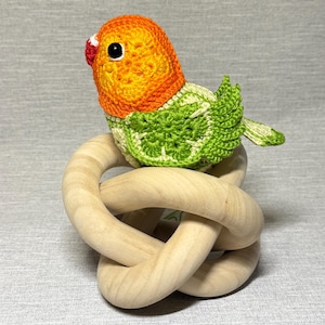 Puede incluir: Un juguete de ganchillo de periquito con plumaje naranja y verde, posado en un anillo de madera anudado.