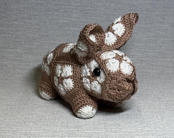 May include: A crochet rabbit stuffed animal constructed from brown and white yarn squares. The rabbit features a repeating floral pattern throughout its body, two long ears, and small black plastic eyes. The paws and face have white accents.