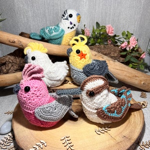 Puede incluir: Cinco pájaros de ganchillo hechos a mano de diferentes colores y especies se exhiben en una rodaja de madera. Los pájaros incluyen un periquito, una cacatúa y otros diseños, mostrando una artesanía detallada. Los pájaros están hechos con hilo.