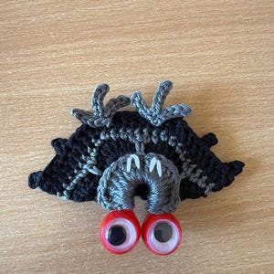 May include: A handmade crochet creature with a black and gray body, gray horns, and large red eyes. The whimsical design is set against a light brown wooden surface. This unique item is perfect for craft enthusiasts.