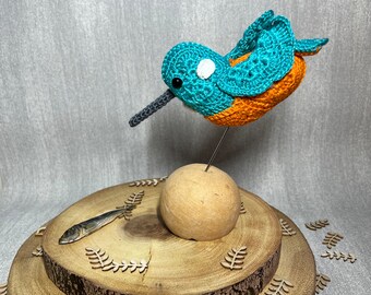 May include: A crocheted blue and orange bird with a white spot on its head, perched on a wooden stand with a small fish beside it.