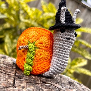 Puede incluir: Un caracol de crochet con un sombrero de bruja negro. La concha del caracol es naranja y verde y se parece a una calabaza.