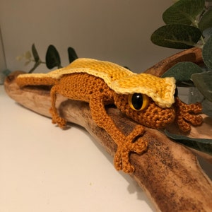 May include: A crocheted gecko toy with a yellow head and orange body. The gecko is sitting on a branch.