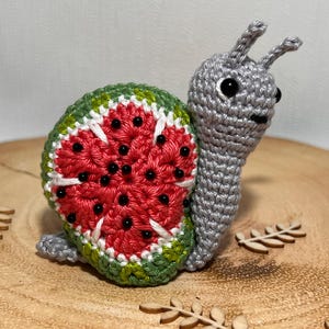 Puede incluir: Un caracol de crochet gris con una concha de sandía. La concha es roja, verde y blanca con semillas negras.