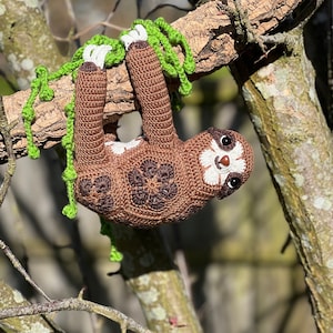 May include: A brown crocheted sloth hanging from a tree branch with green crocheted vines. The sloth has white crocheted details on its face and paws.