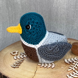 May include: A crocheted duck toy with a teal head, brown and grey body, and a yellow beak. The duck is sitting on a wooden surface with decorative wooden leaves.