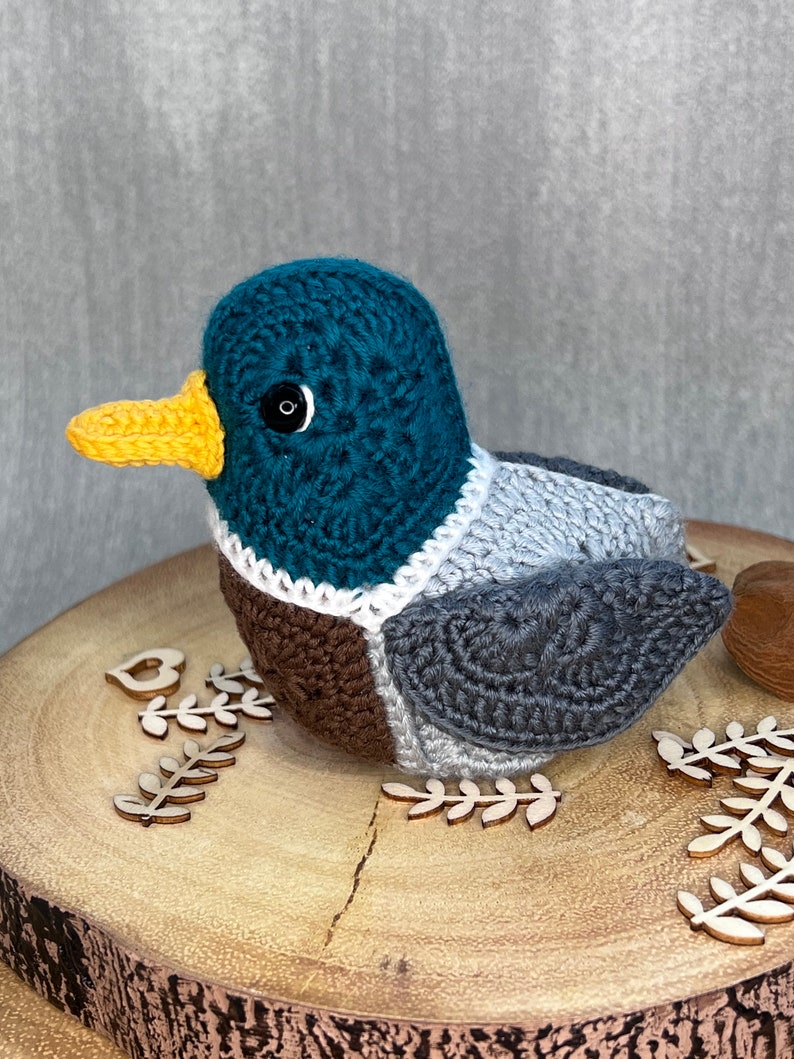 May include: A crocheted duck toy with a teal head, brown and grey body, and a yellow beak. The duck is sitting on a wooden surface with decorative wooden leaves.