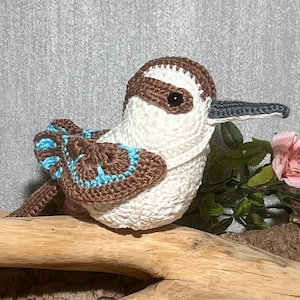 Puede incluir: Un pájaro de peluche tejido a crochet con plumas marrones y blancas y un pico azul. El pájaro está sentado en una rama.