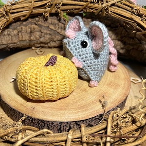 May include: A gray crocheted mouse with pink ears sits next to a yellow crocheted pumpkin on a wooden slice.