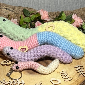 Könnte beinhalten: Vier gehäkelte wurmförmige Schlüsselanhänger in Pastellfarben, darunter rosa, hellblau, Lavendel und grün. Die Schlüsselanhänger sind übereinander gestapelt, wobei der größte unten und der kleinste oben liegt. Jeder Schlüsselanhänger hat einen kleinen Anhänger in Form eines Holzh Herzens.