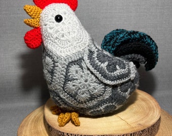 May include: A crocheted gray rooster with a red comb, yellow beak, and teal tail feathers. The rooster is standing on a wooden base.