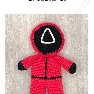 May include: A handmade, red crochet Squid Game Guard doll with black detailing. The doll has a black head with a white triangle, black arms and legs, and the text "Squid Game Guard" at the top.