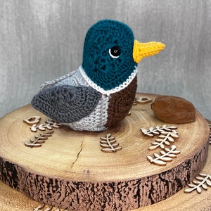 May include: A crocheted duck toy with a teal head, grey body, brown chest, and yellow beak. The duck is sitting on a wooden log with small wooden leaves around it.