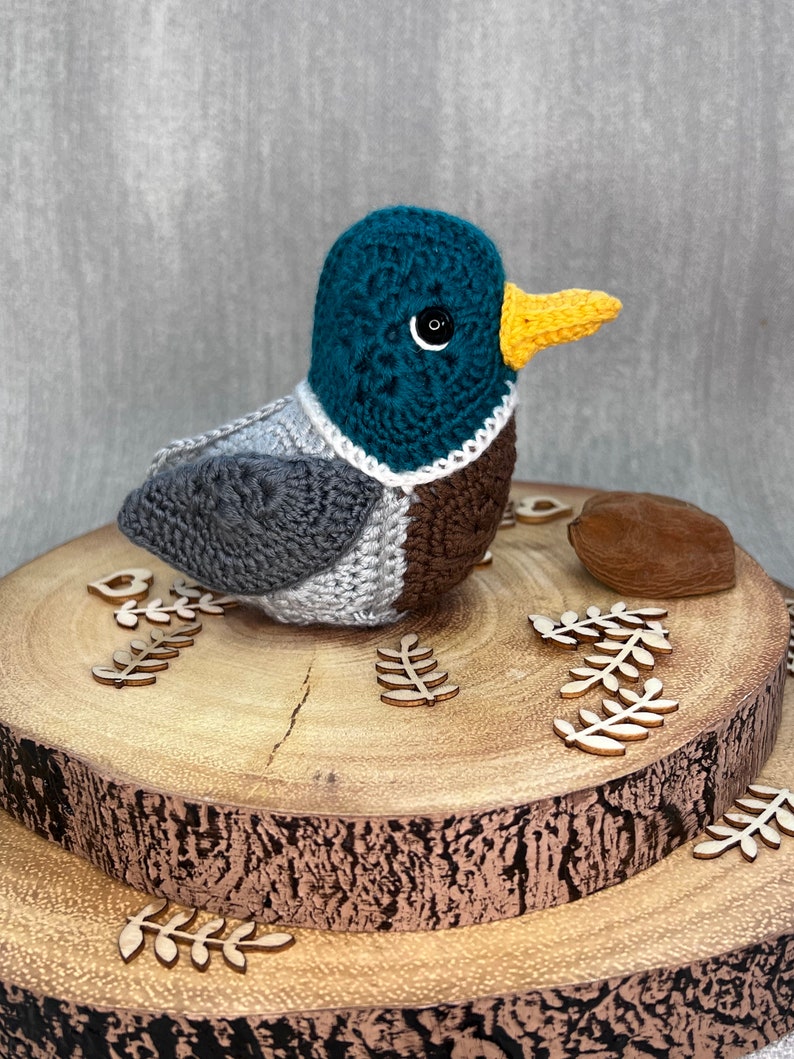 May include: A crocheted duck toy with a teal head, grey body, brown chest, and yellow beak. The duck is sitting on a wooden log with small wooden leaves around it.